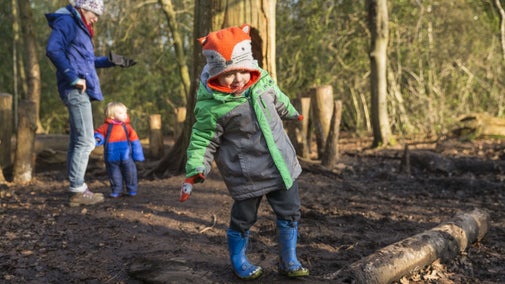 A young child wearing a hat, coat and wellington boots balancing on a log in woodlands